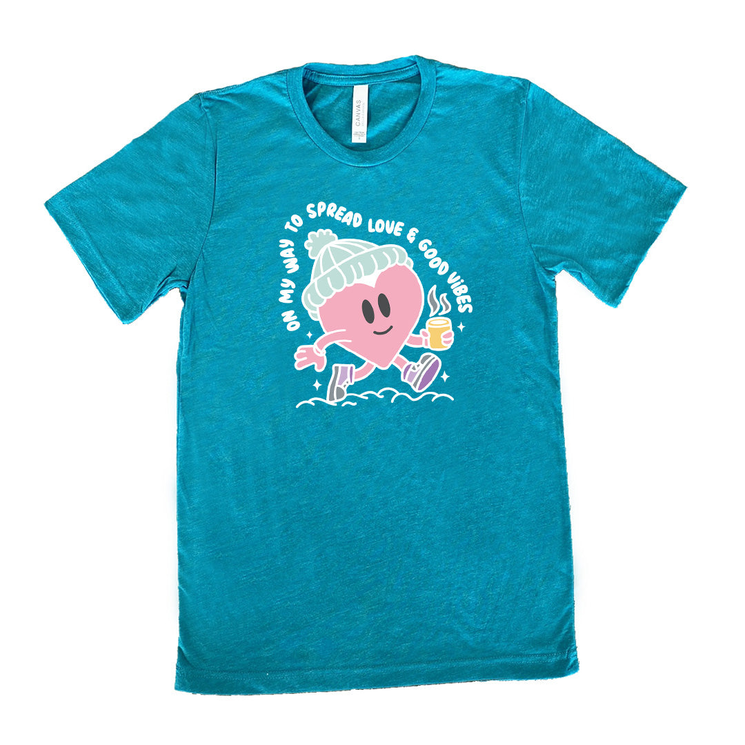 teal unisex tee with the words "on my way to spread love & good vibes" on it, with graphics of a cheerful, cartoony heart strolling along with a mug of a hot drink steaming in its hand, a hat on its head, and shoes on