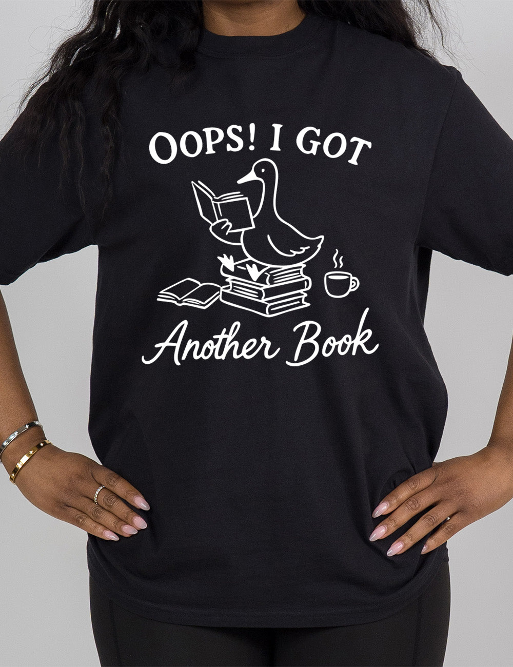 black modern tee with the graphic of a goose, sitting on a stack of books, reading a book, with another book open beside the stack, and a steaming cup of coffee on the other side, with the words around the graphic saying "Oops! I got another book"