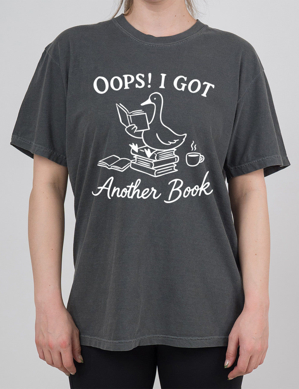 pepper modern tee with the graphic of a goose, sitting on a stack of books, reading a book, with another book open beside the stack, and a steaming cup of coffee on the other side, with the words around the graphic saying "Oops! I got another book"