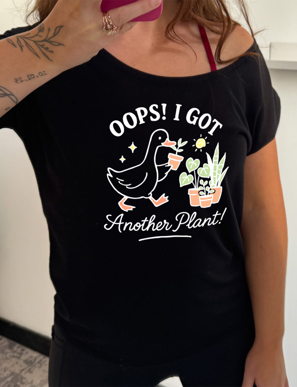 black slouchy tee with the graphic of a goose holding a little plants, walking toward a few other plants, and the words surrounding it saying "Oops! I got another plant!"