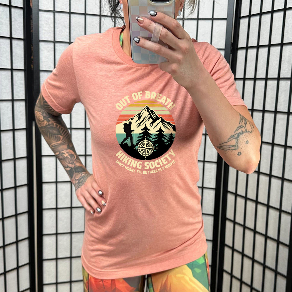 Sunset colored shirt with a hiker and mountain graphic on it that says "Out Of Breath Hiking Society"