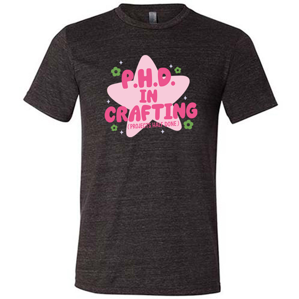 black unisex tee with cartoon graphic of a large star behind the words "P.H.D. in Crafting" in large font with the words (Projects Half Done) in small font underneath, with very small flowers and stars around