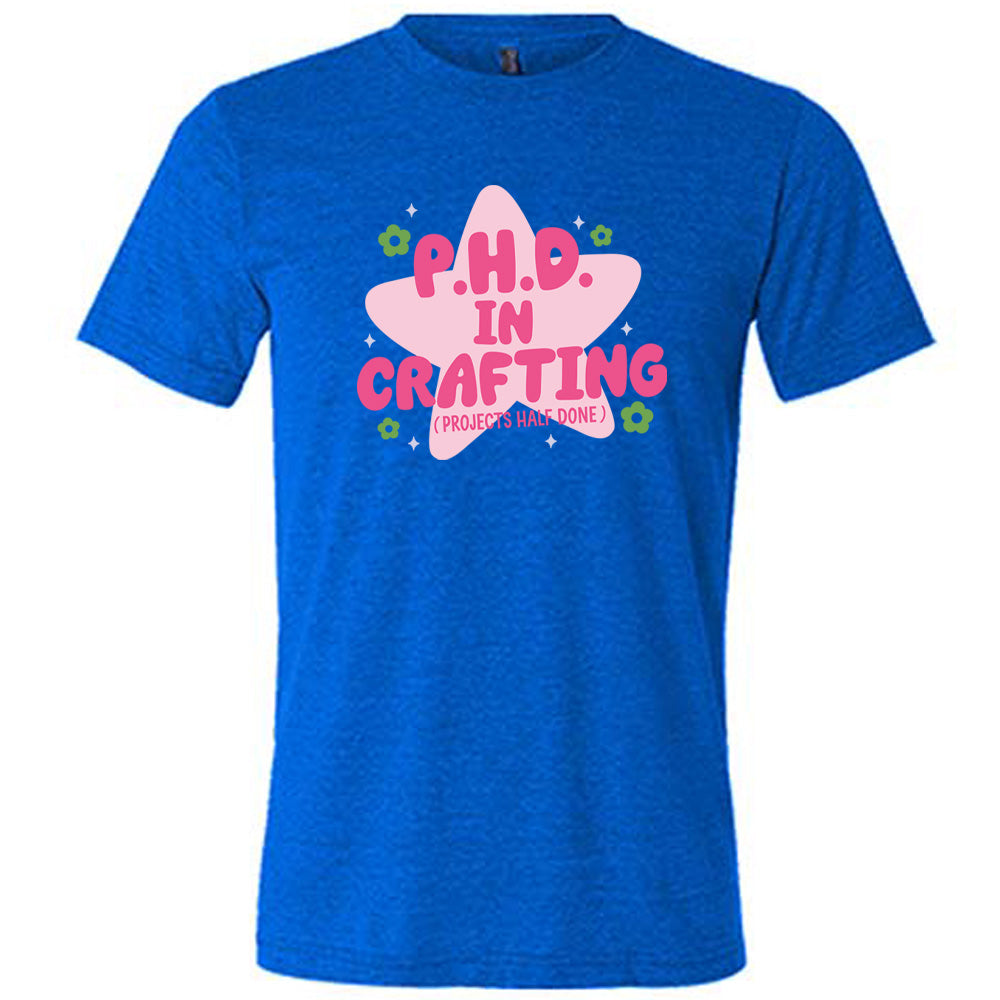 blue unisex tee with cartoon graphic of a large star behind the words "P.H.D. in Crafting" in large font with the words (Projects Half Done) in small font underneath, with very small flowers and stars around