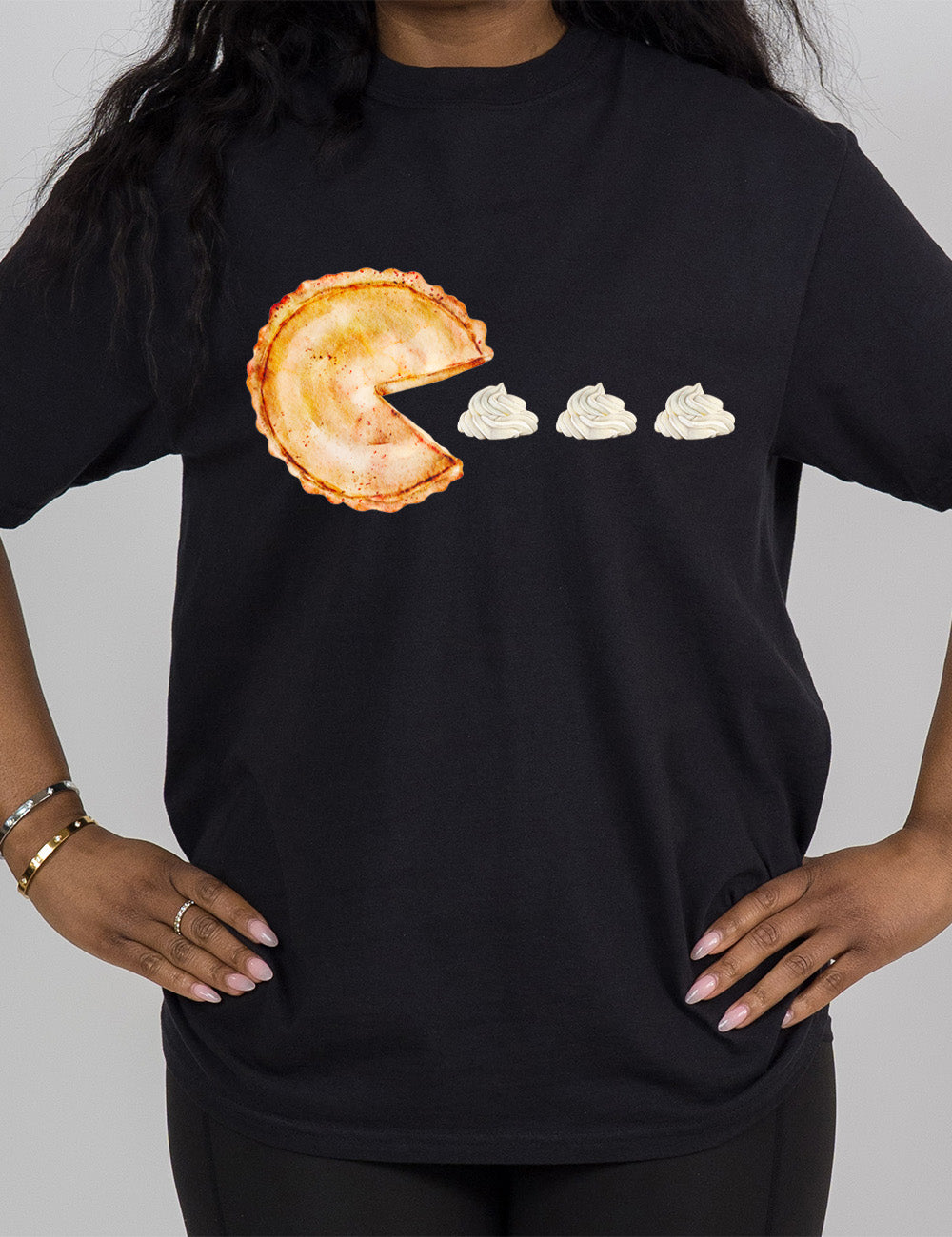 black modern tee with the graphic of a pumpkin pie, with the a slice taken out of it so that it looks like Pacman, heading in the direction of three dollops of whipped cream