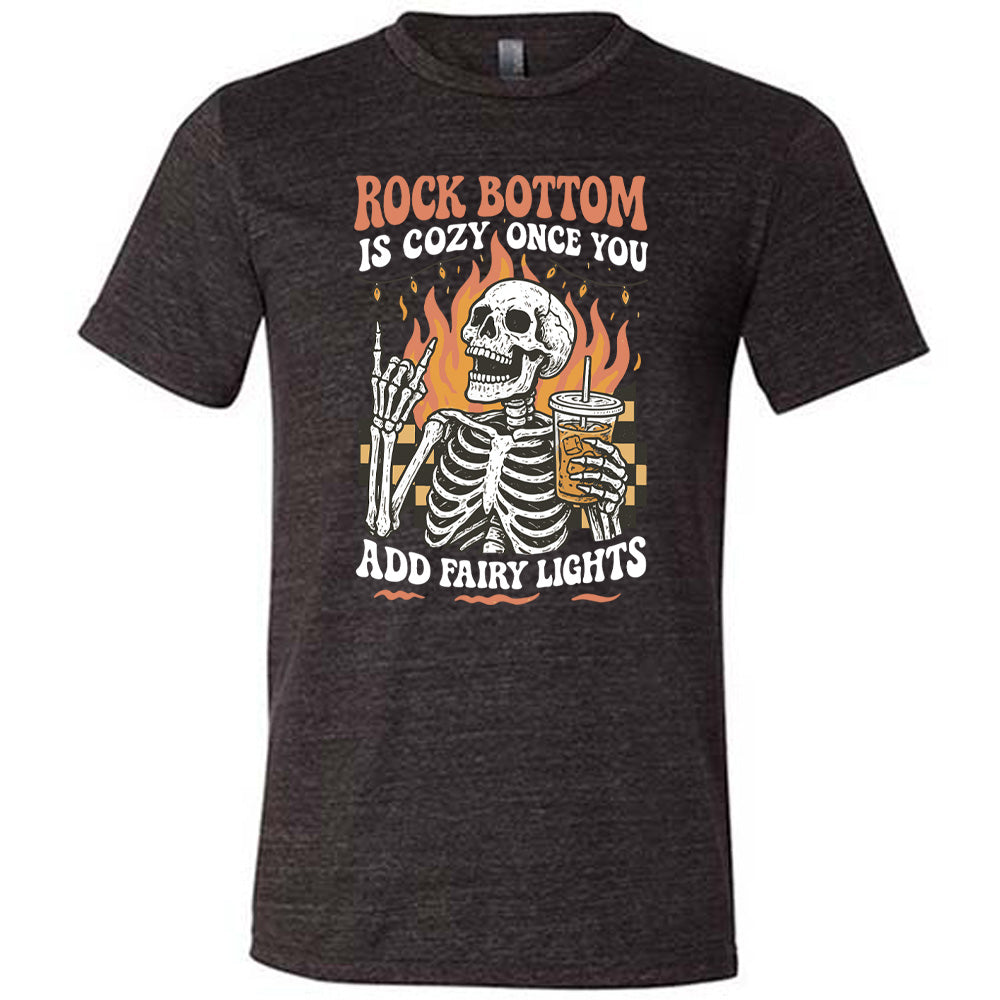 black unisex tee with the words "rock bottom is cozy once you add fairy lights" with the graphic of a skeleton giving the rocker sign with one hand while holding an iced drink in the other, with a background of some checkerboard, some flames, and a string of fairy lights above the flames