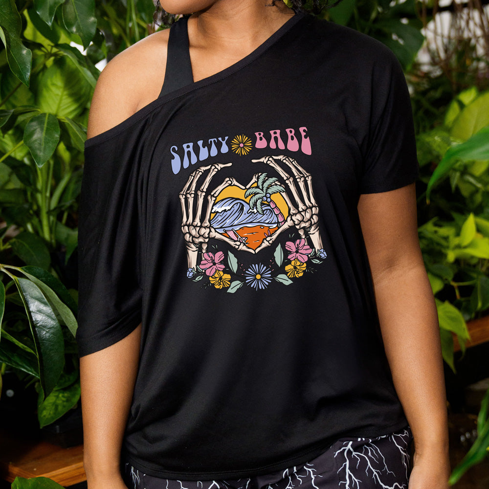 Black colored shirt with a skeleton heart hands around ocean waves graphic on it that says "Salty Babe"
