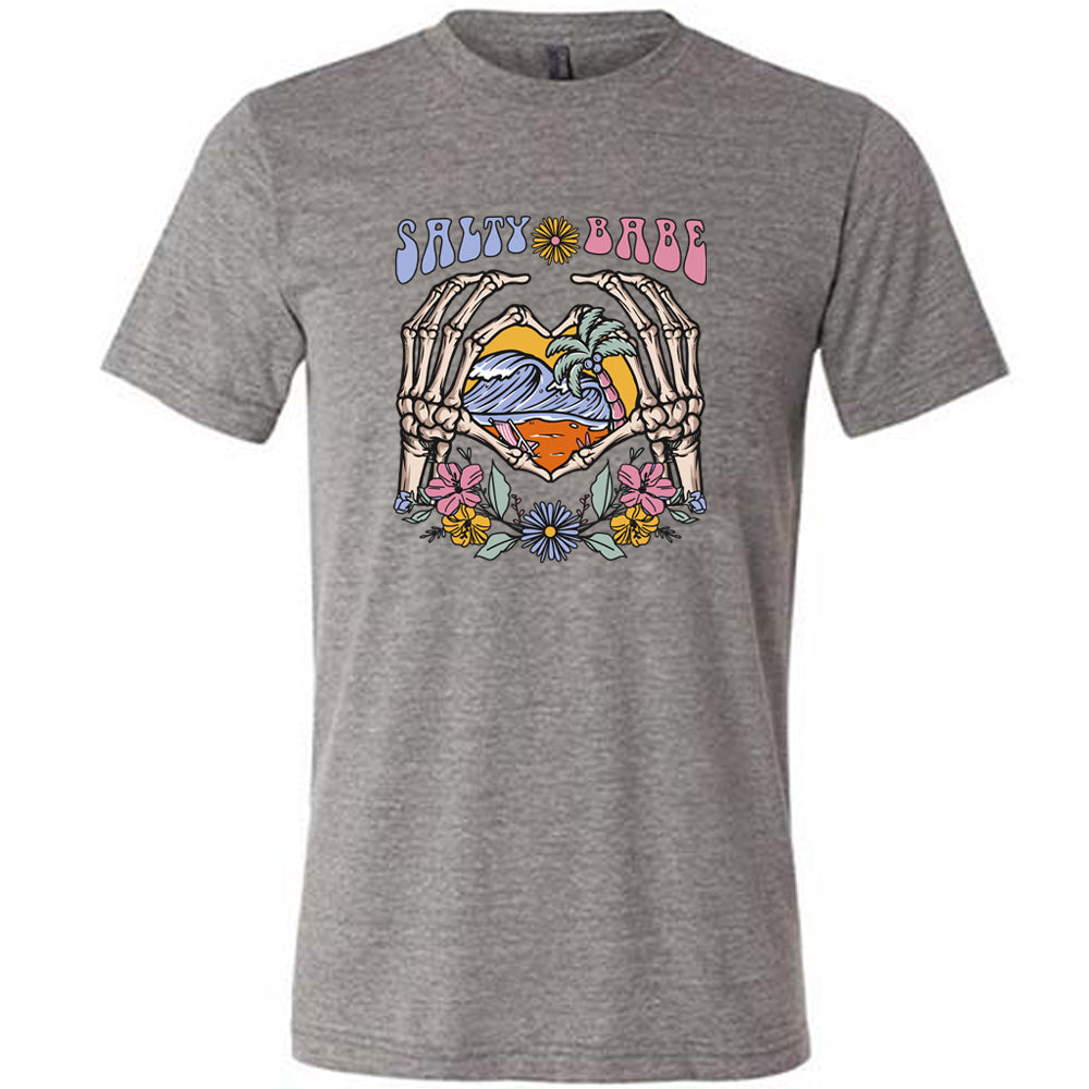 Grey colored shirt with a skeleton heart hands around ocean waves graphic on it that says "Salty Babe"