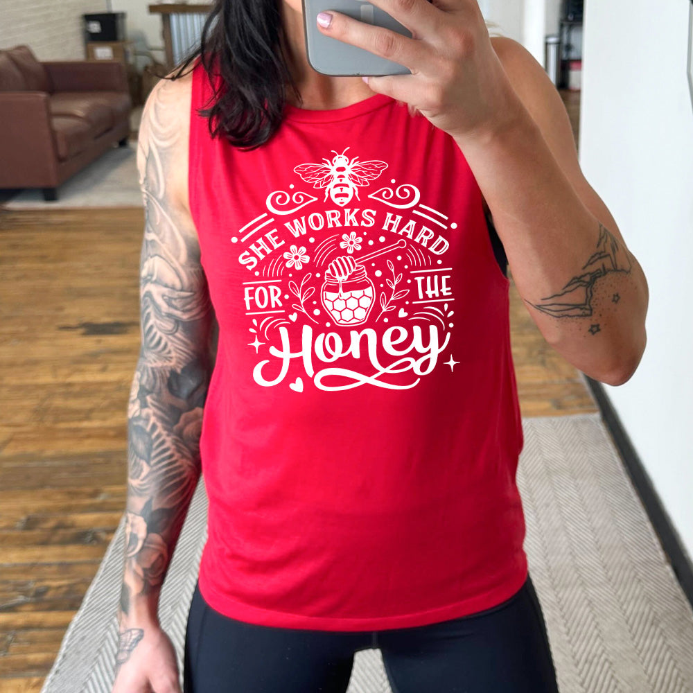red muscle tank with the words "she works for the honey" surrounded by accenting curved lines, with flowers, dots, stars, and hears scattered around, and a honeypot and spoon in the center, and a bee on the top