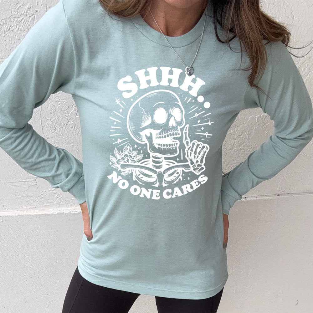 heather dusty blue long sleeve with the words "Shhh.. no one cares" on it, with the graphic of a skeleton holding a finger to its lips