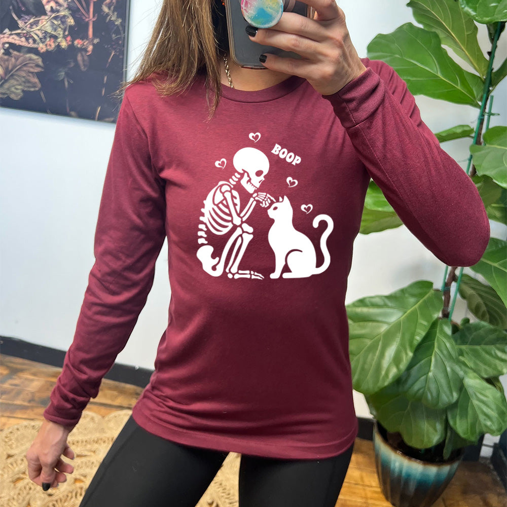 heather cardinal long sleeve shirt with the graphic of a sitting skeleton touching the head of a sitting cat, with the word "boop" above this action, and four little hearts around them