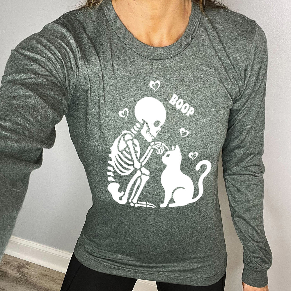 heather forest long sleeve shirt with the graphic of a sitting skeleton touching the head of a sitting cat, with the word "boop" above this action, and four little hearts around them