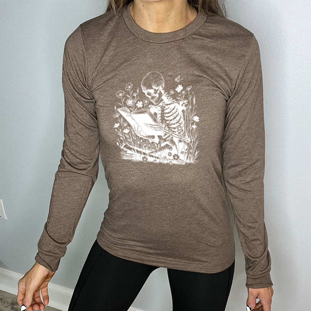 heather brown long sleeve shirt with the graphic of a skeleton reading a book, sitting cross-legged in a bed of tall flowers