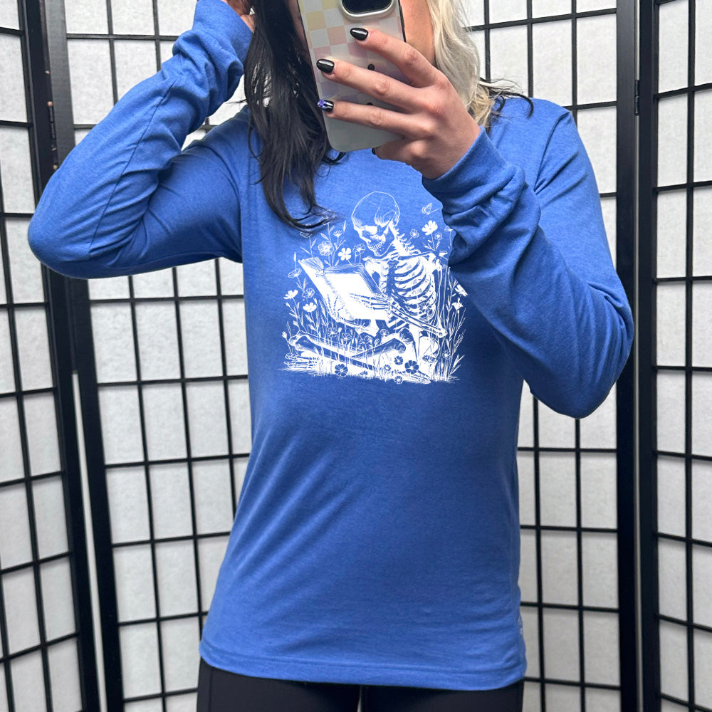 heather true royal long sleeve shirt with the graphic of a skeleton reading a book, sitting cross-legged in a bed of tall flowers