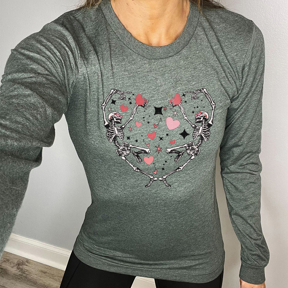 heather forest long sleeve shirt with the graphic of two skeletons creating the shape of a heart with their bodies, appearing to be dancing, with many hearts between them, and a few little stars