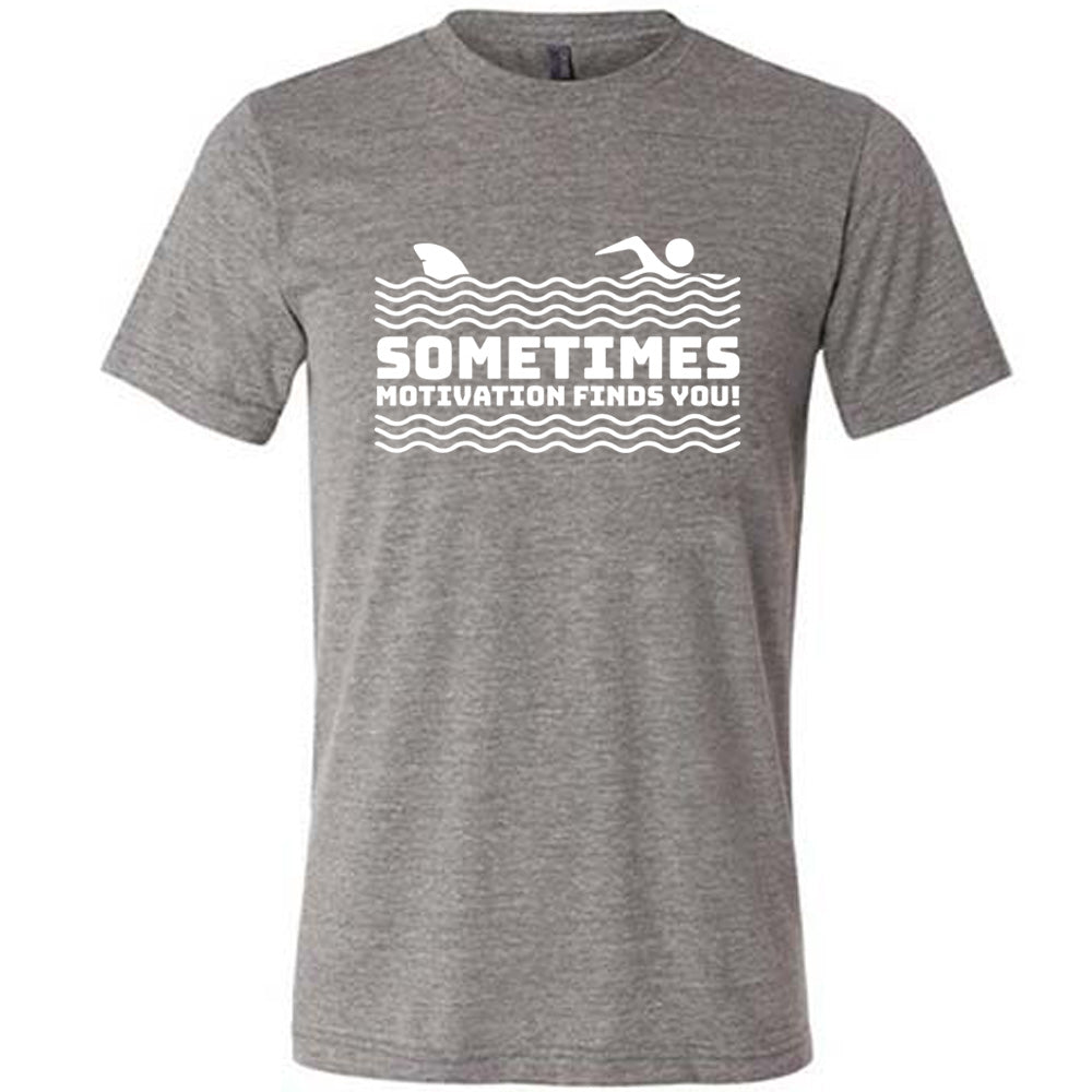 grey unisex tee with the words "sometimes motivation finds you!" in between the lined graphic of waves of the sea, and on top of the waves is the graphic of a figure swimming away from a fin in the water behind