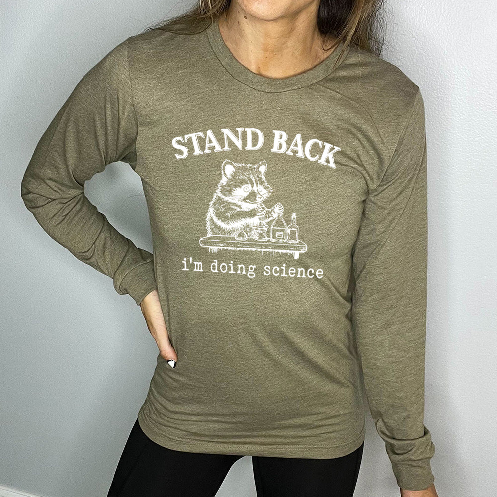 heather olive long sleeve shirt with the words "stand back i'm doing science" on it, with the graphic of a racoon with its paws raised over various bottles