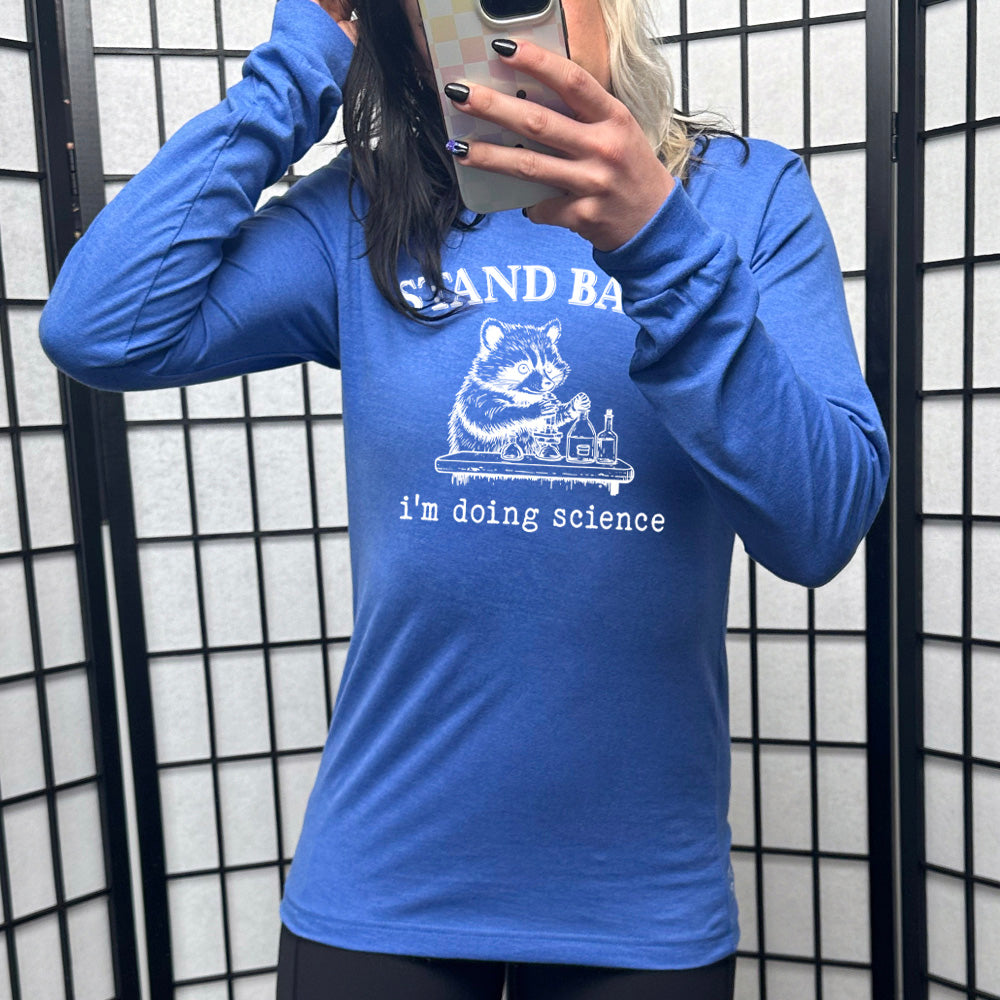 heather true royal long sleeve shirt with the words "stand back i'm doing science" on it, with the graphic of a racoon with its paws raised over various bottles