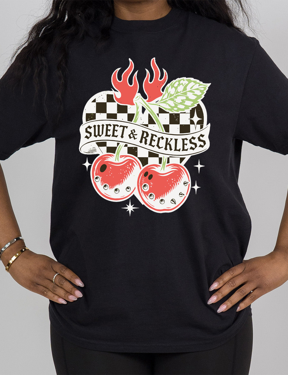black modern tee with print placement on the front, with the graphic of two large cherries, with spikes, on connected stems with a leaf, in front of a checkered heart, with flames on top, a few stars around, with a banner across the heart saying "sweet & reckless"