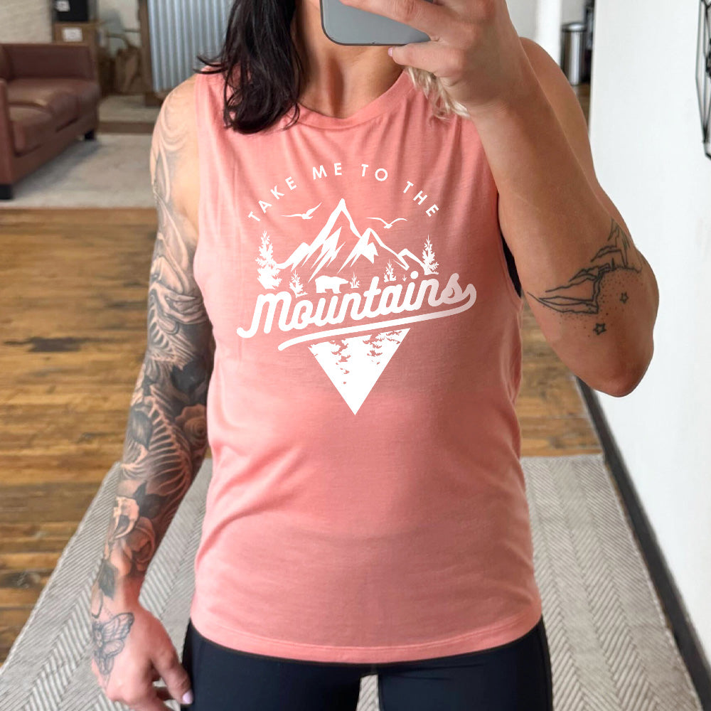 Model wearing peach colored muscle tank with a mountains graphic on it that says "Take Me To The Mountains"