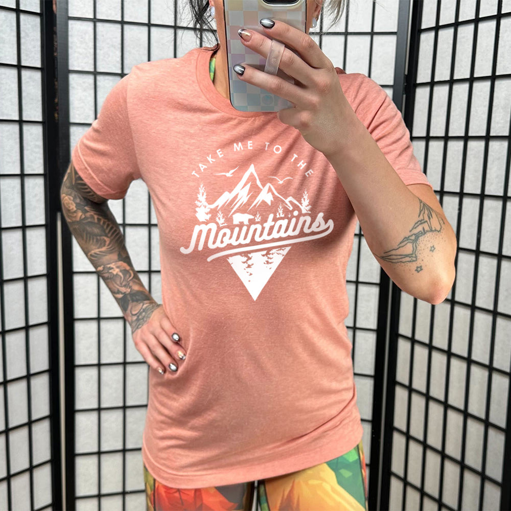 Model wearing sunset colored shirt with a mountains graphic on it that says "Take Me To The Mountains"