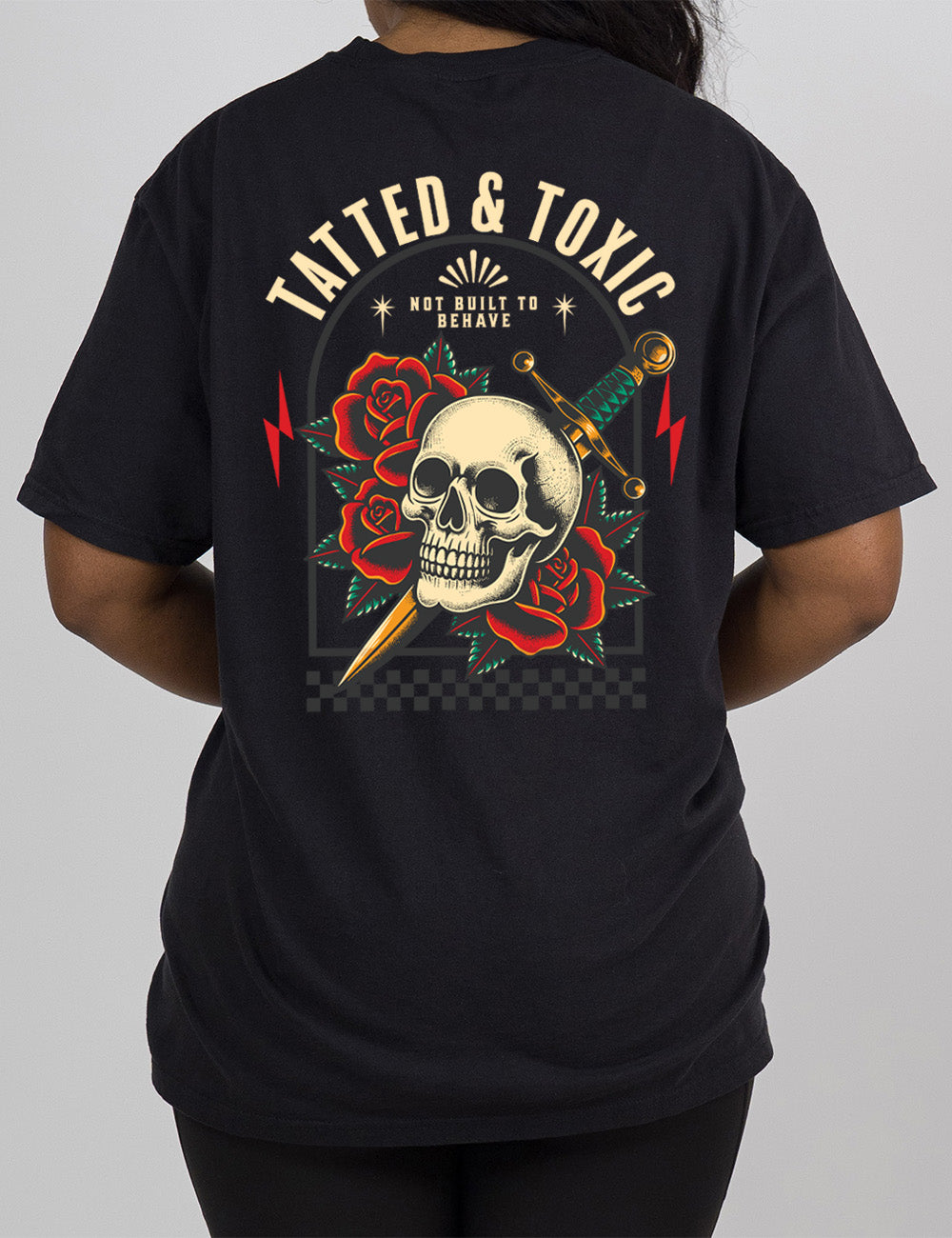 black modern tee with the print placement being on the back, with the words "tatted & toxic" on it, with the graphic of a skull, with a knife through its head, among roses, with small words above it, saying "not built to behave," within an arched box, over a strip of checkered design, with a lightning bolt on either side