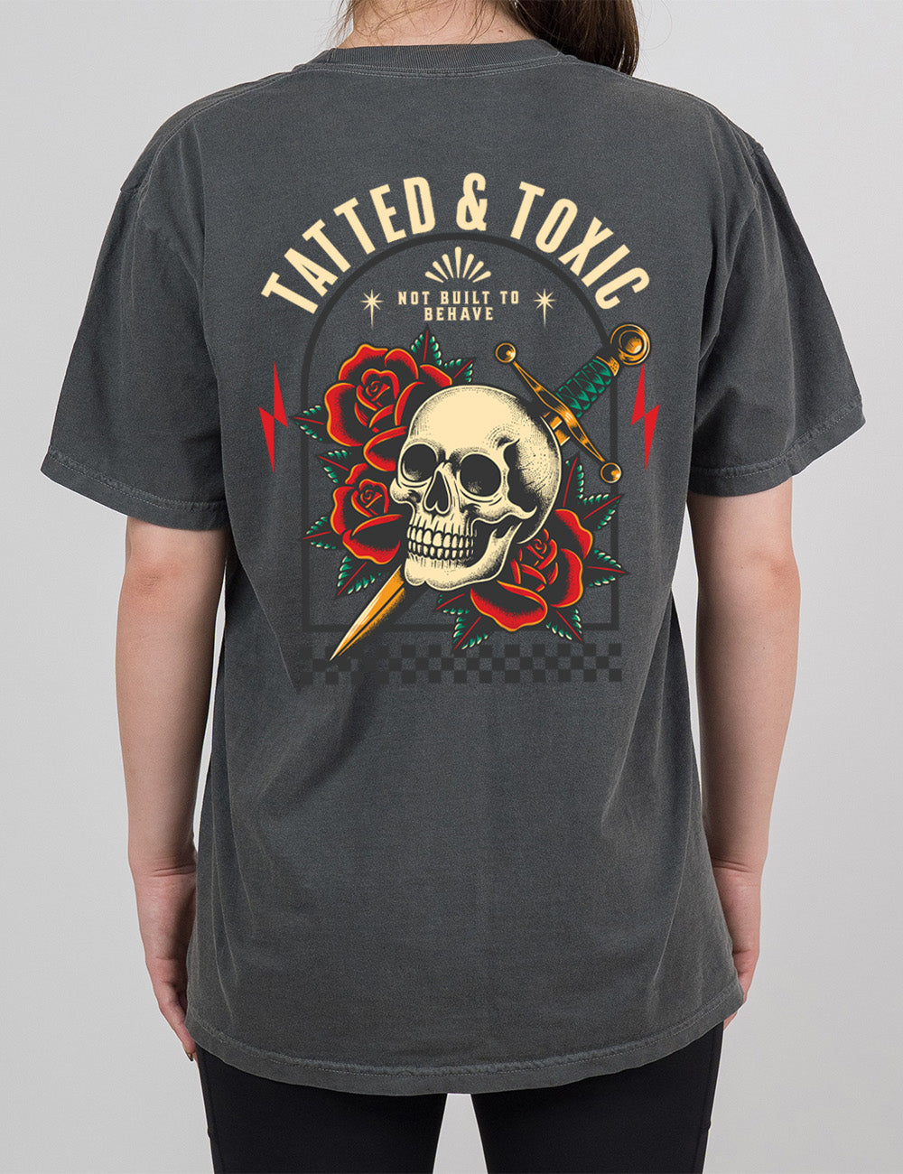 pepper modern tee with the print placement being on the back, with the words "tatted & toxic" on it, with the graphic of a skull, with a knife through its head, among roses, with small words above it, saying "not built to behave," within an arched box, over a strip of checkered design, with a lightning bolt on either side