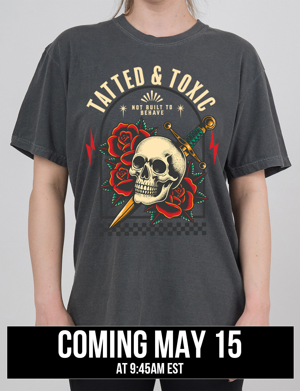 pepper modern tee with the print placement being on the front, with the words "tatted & toxic" on it, with the graphic of a skull, with a knife through its head, among roses, with small words above it, saying "not built to behave," within an arched box, over a strip of checkered design, with a lightning bolt on either side, coming may 15th