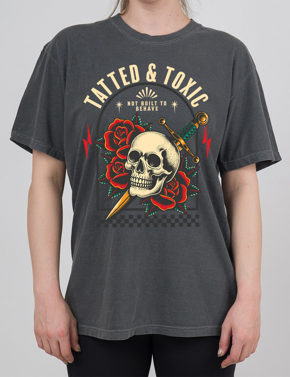pepper modern tee with the print placement being on the front, with the words "tatted & toxic" on it, with the graphic of a skull, with a knife through its head, among roses, with small words above it, saying "not built to behave," within an arched box, over a strip of checkered design, with a lightning bolt on either side