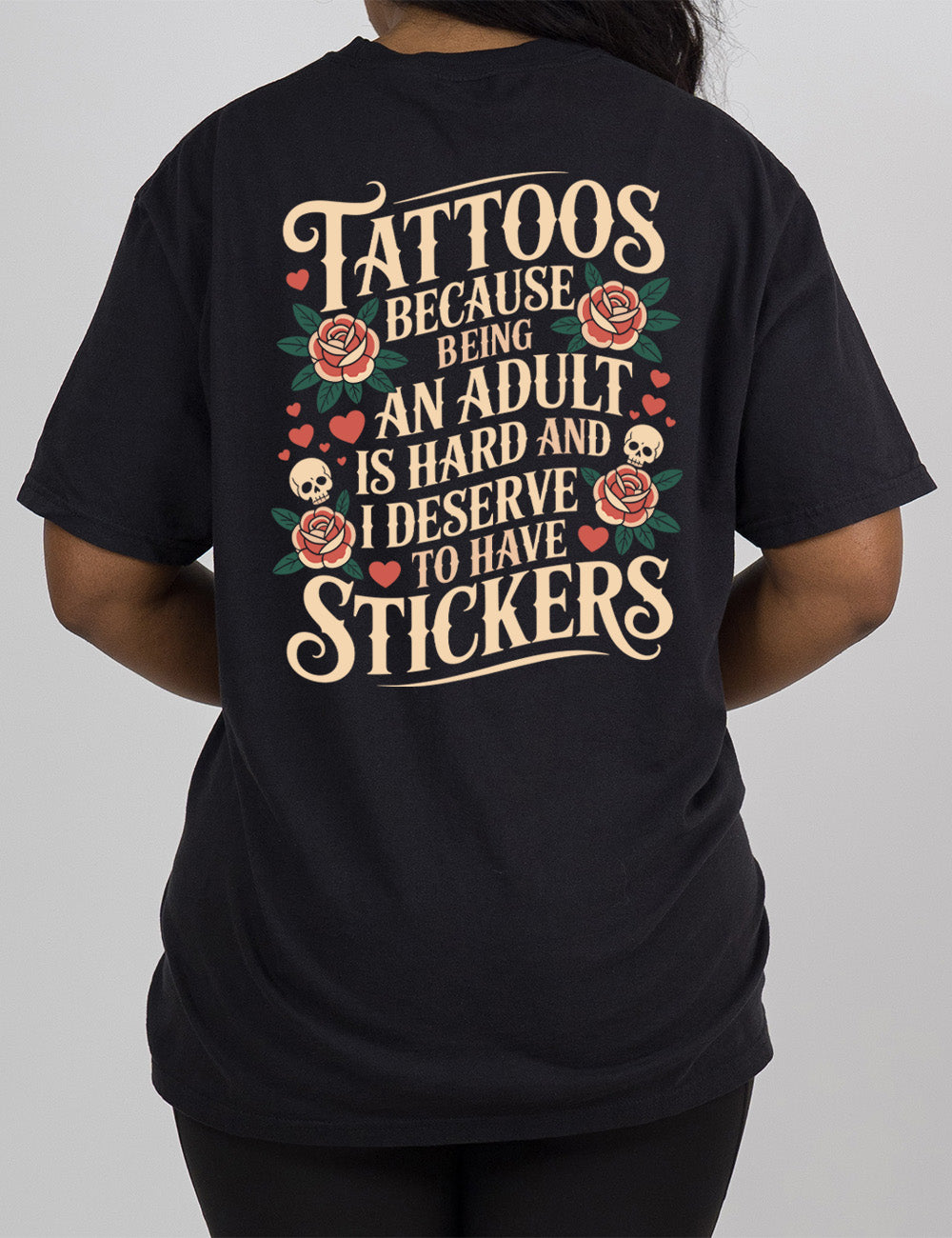 black modern tee with print placement on the back, with the words "tattoos because being an adult is hard and I deserve to have stickers" on it, with the graphics of roses, hearts, and skulls on the side of the words