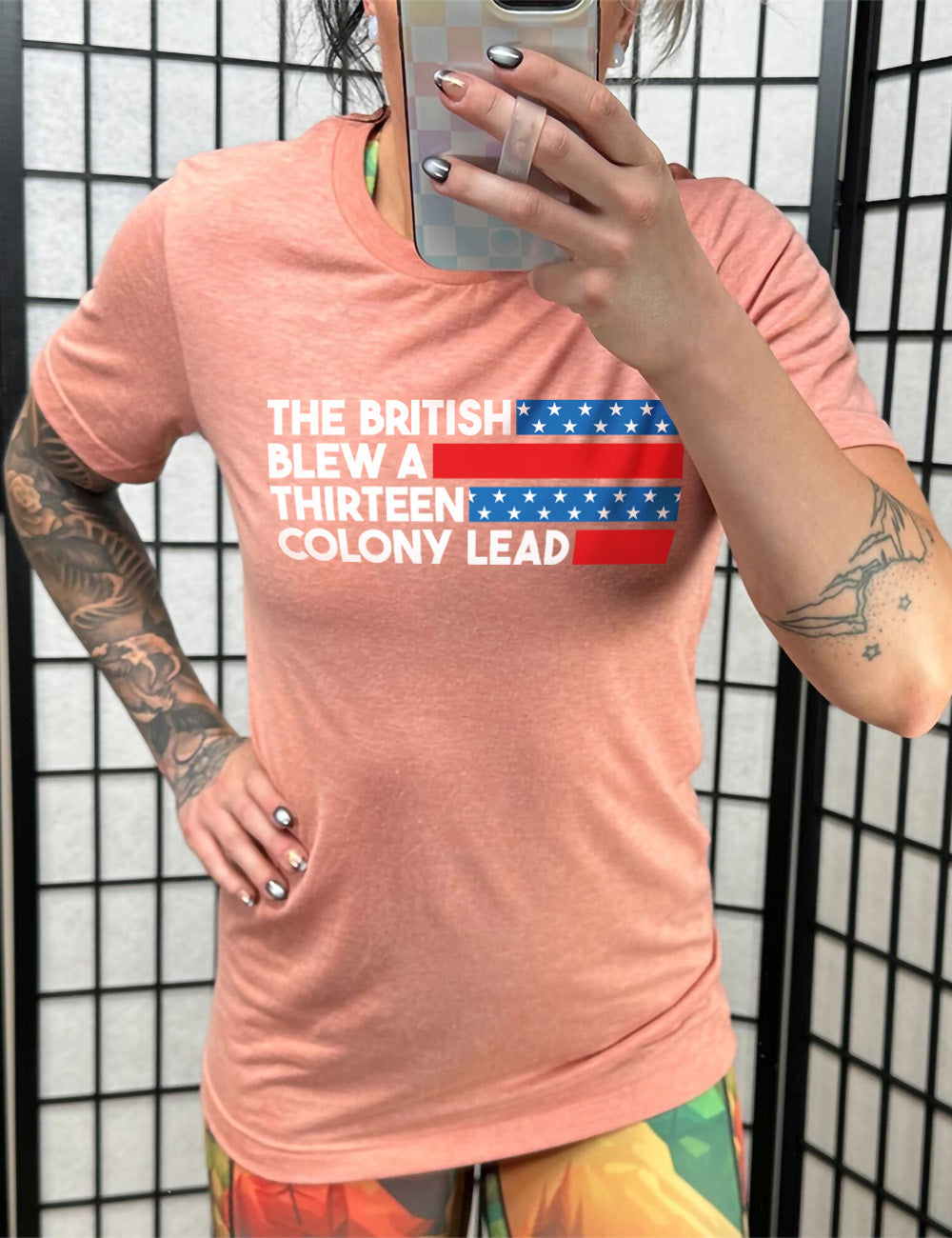 sunset unisex tee with the words "the British" (and a blue starred bar finishing the space to the right), then below "blew a" (and a red bar), then below "thirteen" (and a blue starred bar), and below "colony lead" (and a red bar)