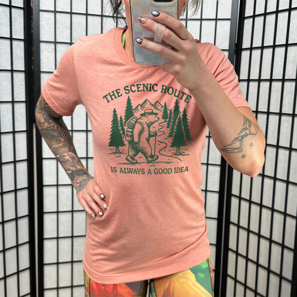 sunset unisex tee with the words "the scenic route is always a good idea" with a bear hiking on a trail through the woods