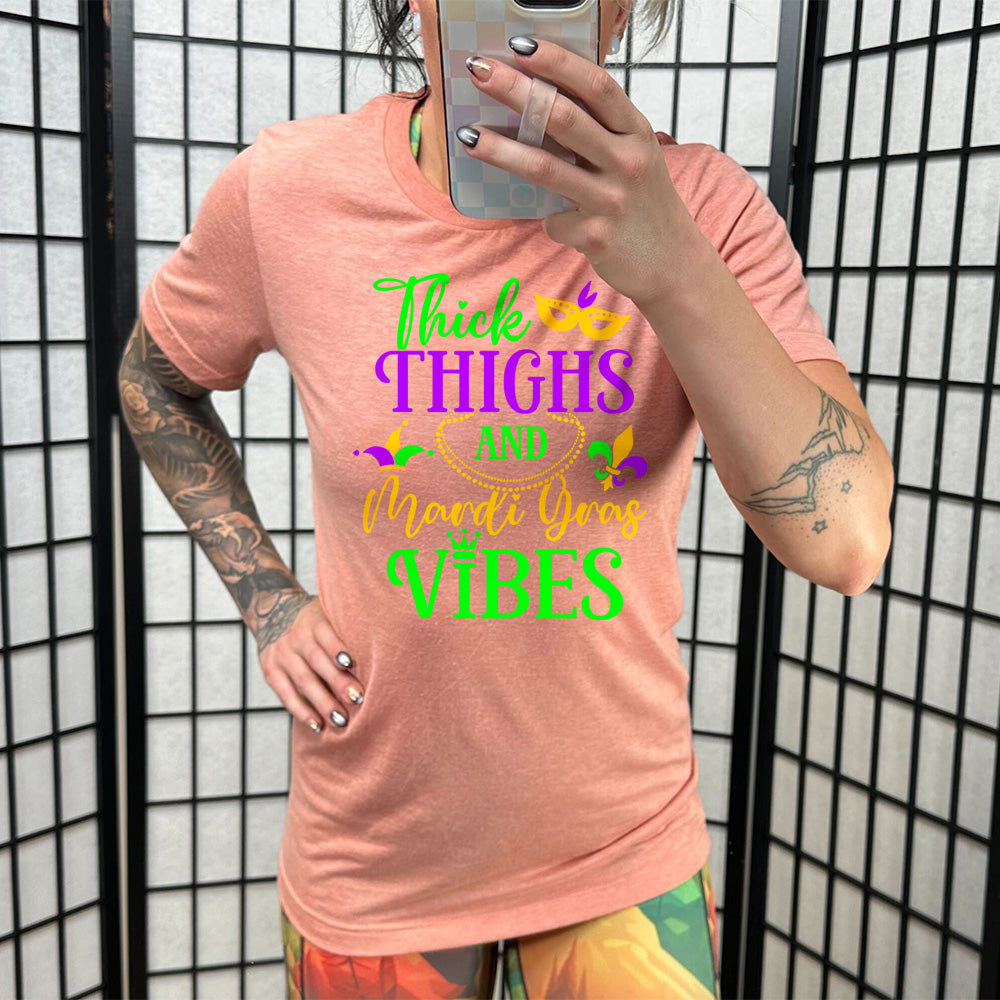 sunset unisex tee with the words "thick thighs and mardi gras vibes" on it in varying font, with small graphics of mardi gras beads, fleur de lis, a joker hat and a mask