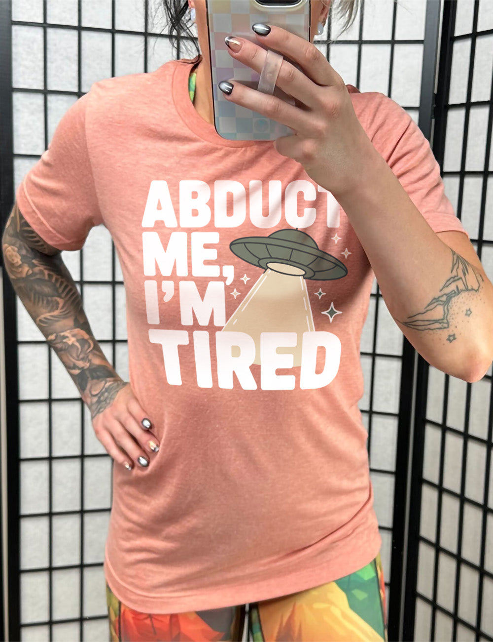 sunset unisex tee with the words "abduct me, I'm tired" on it, with a graphic of an alien spaceship, with a beam extended, and a few little stars