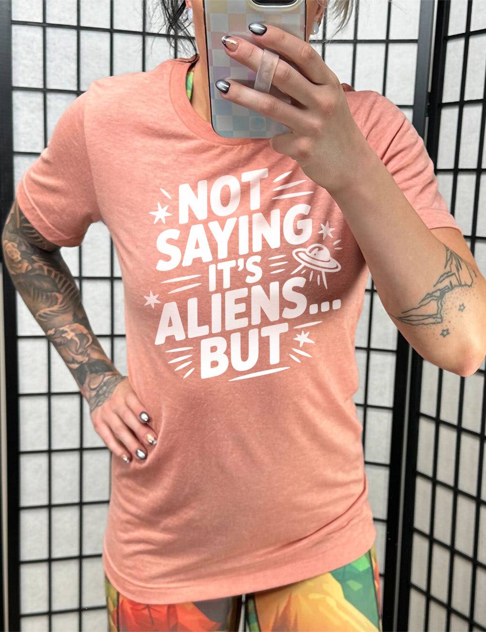 sunset unisex tee with words, embellished by line graphics, saying "not saying it's aliens...but" with graphics of little stars and an alien spaceship