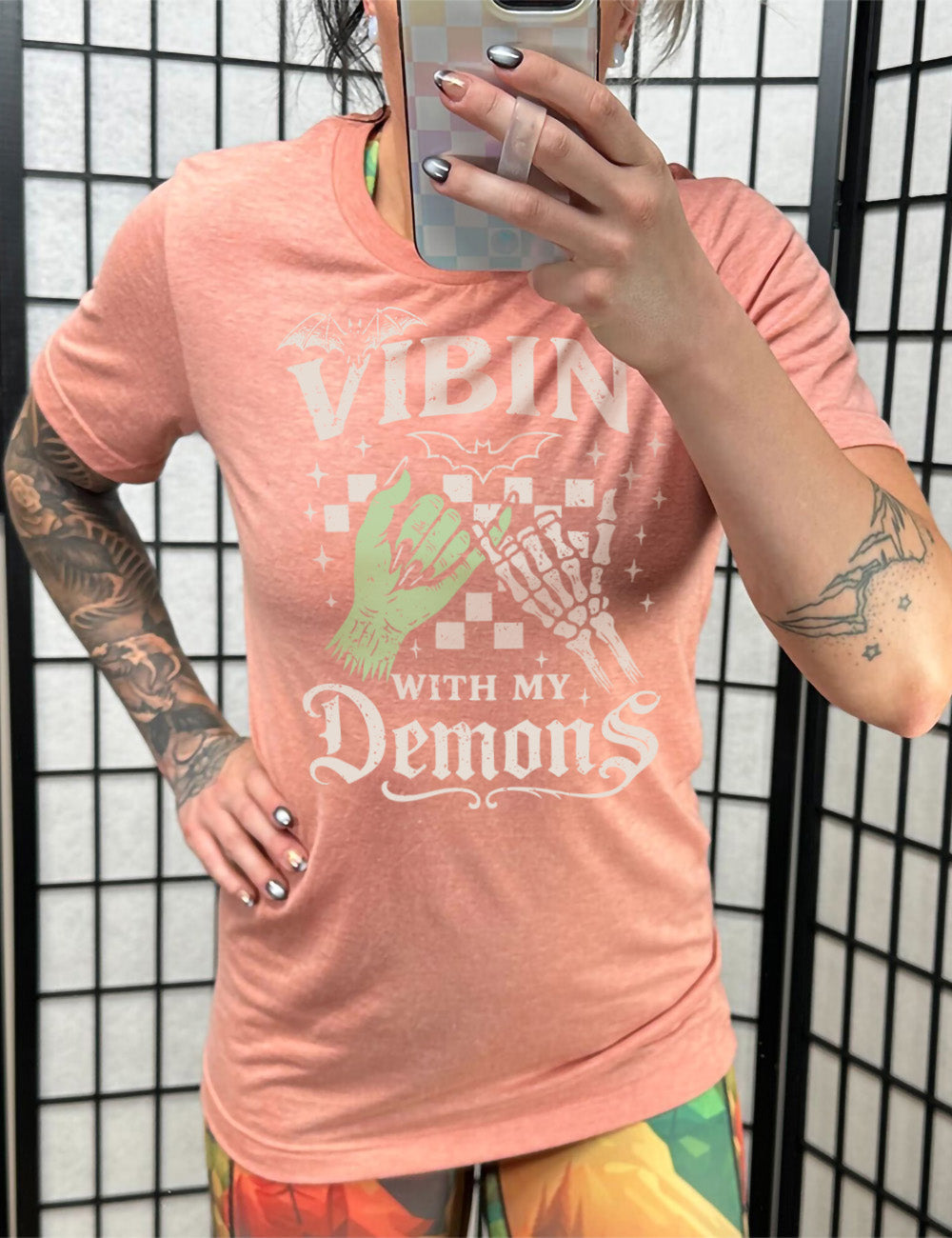 sunset unisex tee with words saying "Vibin' with my Demons", around the graphic of the hands of a zombie and a skeleton doing a pinky promise, in front of a checkered background, and one line-drawn bat and one detailed bat on top, and scattered stars