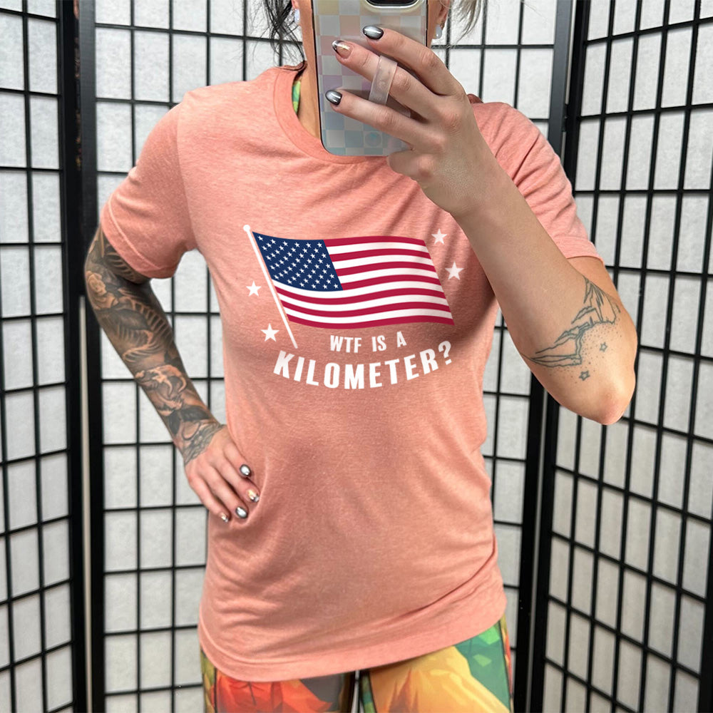 Model wearing sunset colored shirt with an American flag graphic on it that says "WTF Is A Kilometer?"