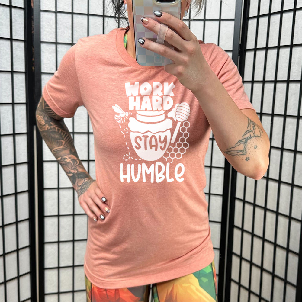 sunset unisex tee with the words "work hard stay humble" on it, with the word "stay" on the graphic of a honeypot, and a bee flying around the pot's left, and some honeycomb and a honey spoon on its right