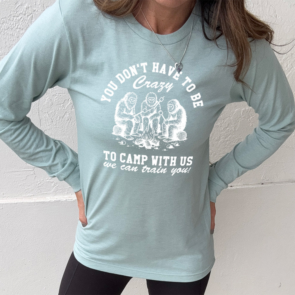 heather dusty blue long sleeve with the words "you don't have to be crazy to camp with us we can train you!" on it with the graphic of three sasquatches toasting marshmallows over a campfire