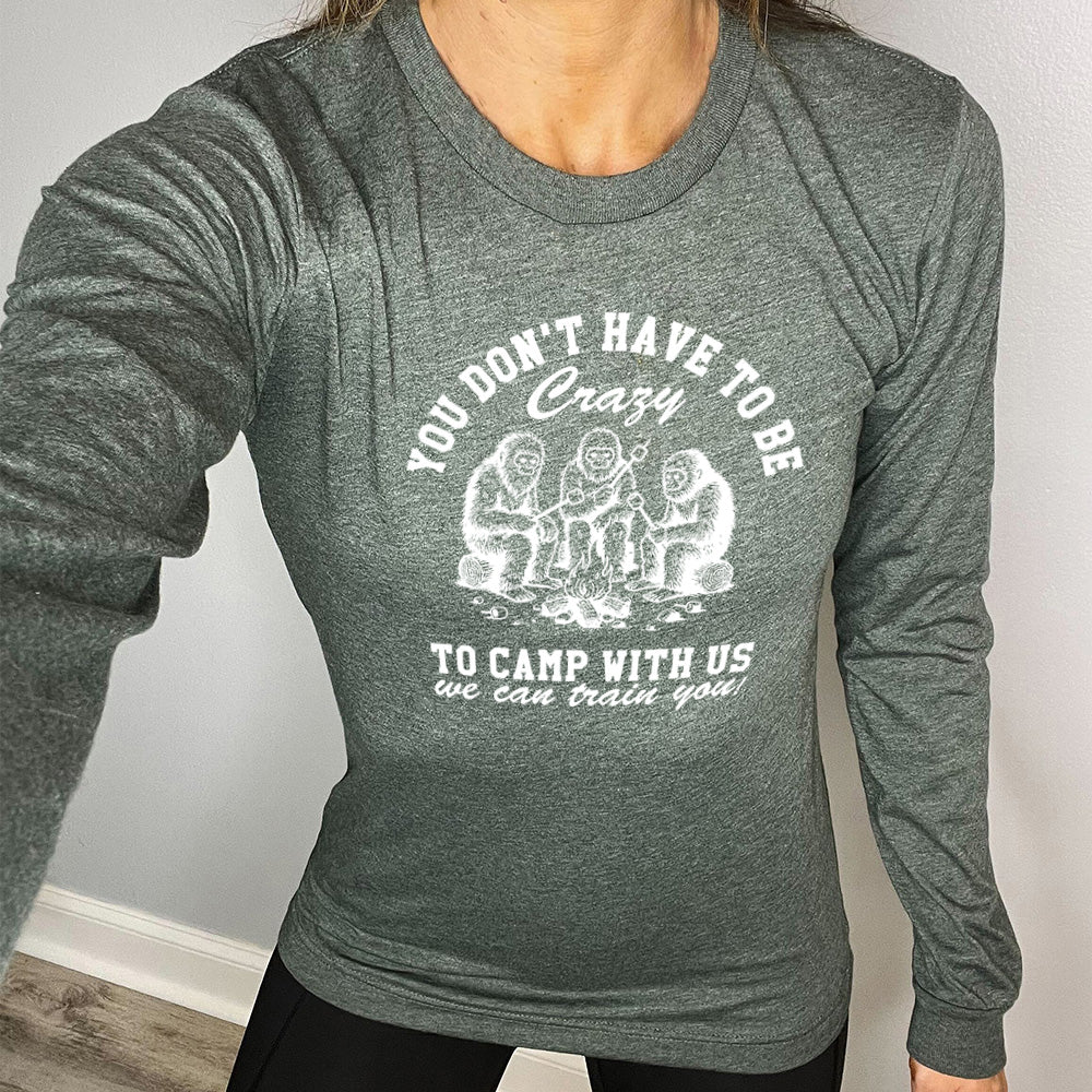 heather forest long sleeve with the words "you don't have to be crazy to camp with us we can train you!" on it with the graphic of three sasquatches toasting marshmallows over a campfire