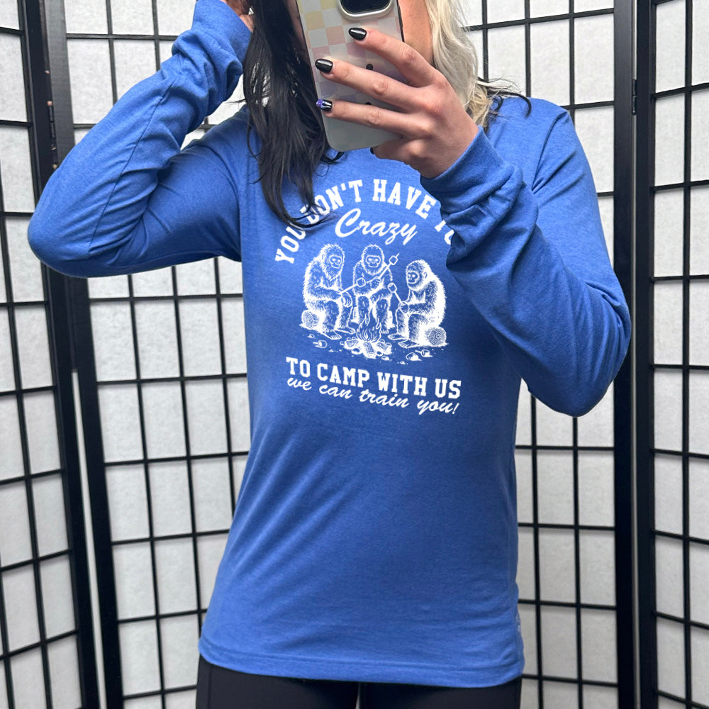 heather true royal long sleeve with the words "you don't have to be crazy to camp with us we can train you!" on it with the graphic of three sasquatches toasting marshmallows over a campfire