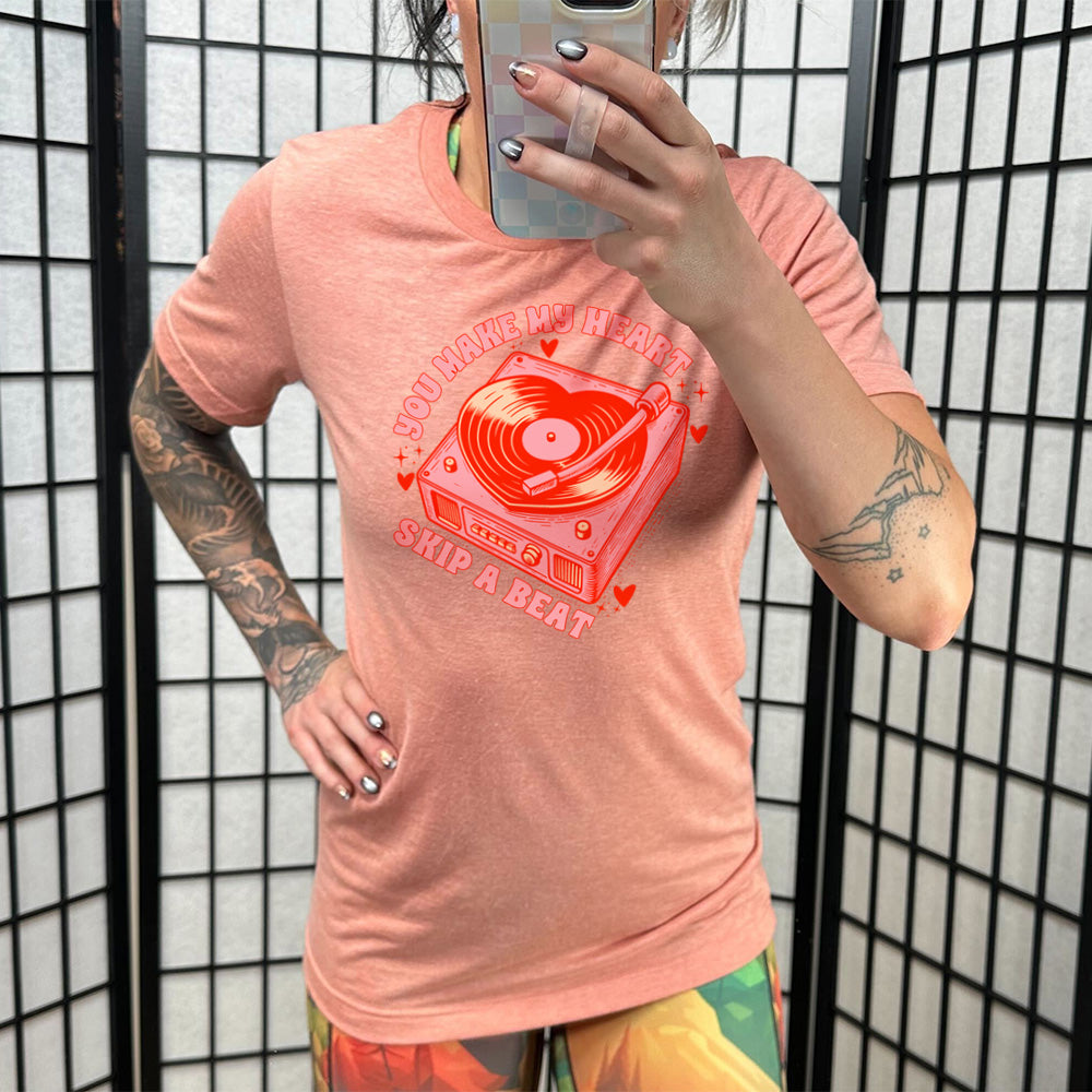 sunset unisex tee with the words "you make my heart skip a beat" on it, with the graphics of a record player, with a record shaped like a heart, with a little hearts and stars around each corner of the player