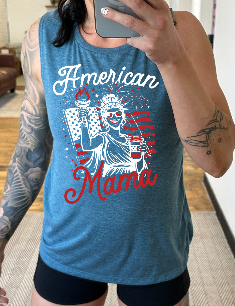 deep teal muscle tank with the graphic of the statue of liberty in front of an American flag, fireworks above, wearing American flag sunglasses, holding a American flag coffee cup, with the words "American Mama" surrounding it