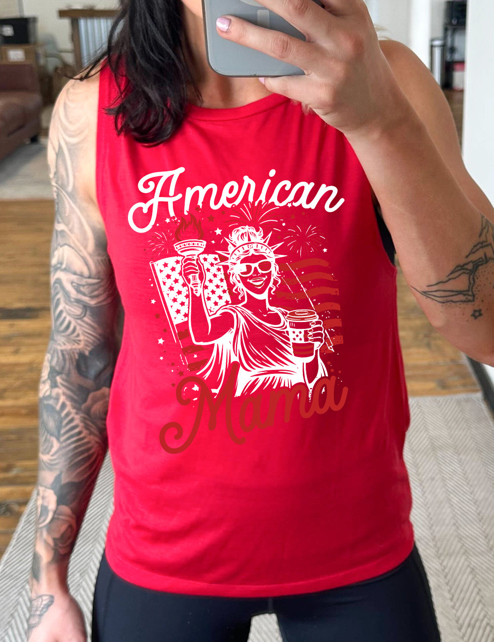 red muscle tank with the graphic of the statue of liberty in front of an American flag, fireworks above, wearing American flag sunglasses, holding a American flag coffee cup, with the words "American Mama" surrounding it
