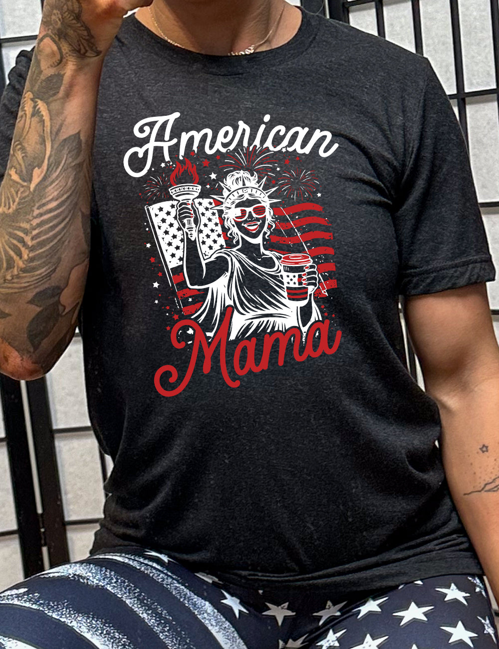black unisex tee with the graphic of the statue of liberty in front of an American flag, fireworks above, wearing American flag sunglasses, holding a American flag coffee cup, with the words "American Mama" surrounding it