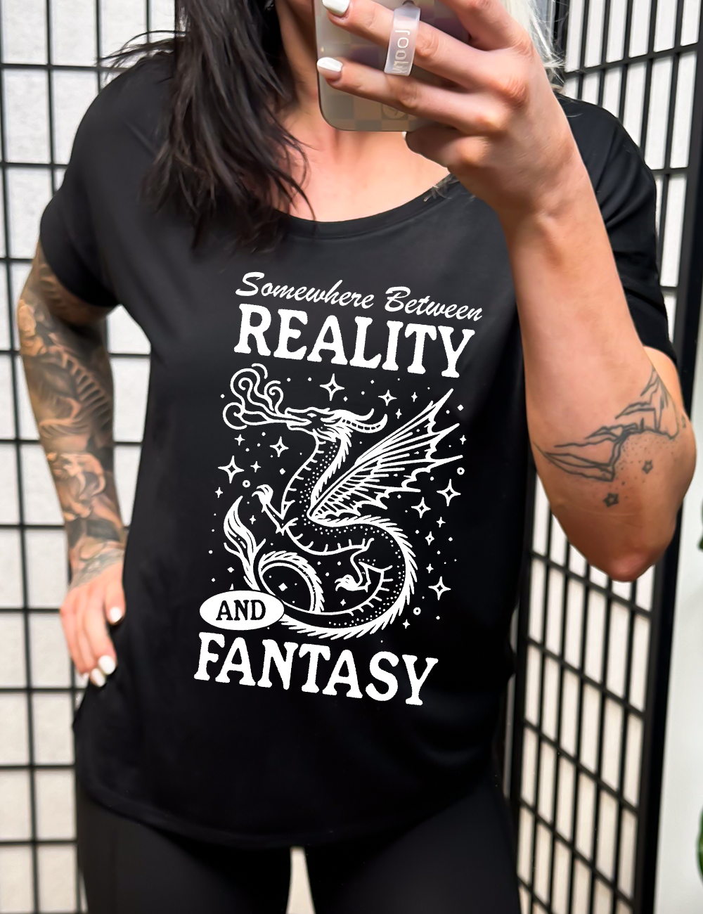 black slouchy tee with the words "somewhere between reality and fantasy" on it, around the graphic of a dragon among stars, breathing fire