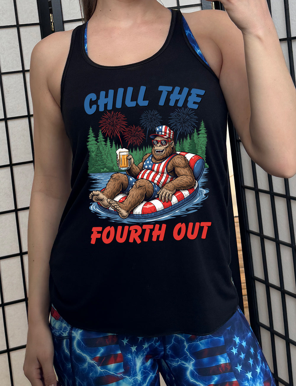 black flowy racerback tank with the graphic of a grinning Bigfoot holding a beer, wearing sunglasses and an American flag outfit, in an American flag tube, in the water, with a background of trees, with fireworks above, and the words saying "chill the fourth out" on it