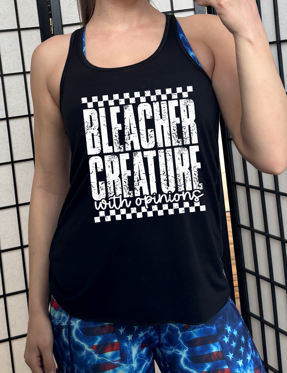 black flowy racerback tank with the large words "bleacher creature" and the small words underneath "with opinions", all of which has a top and bottom checkered border