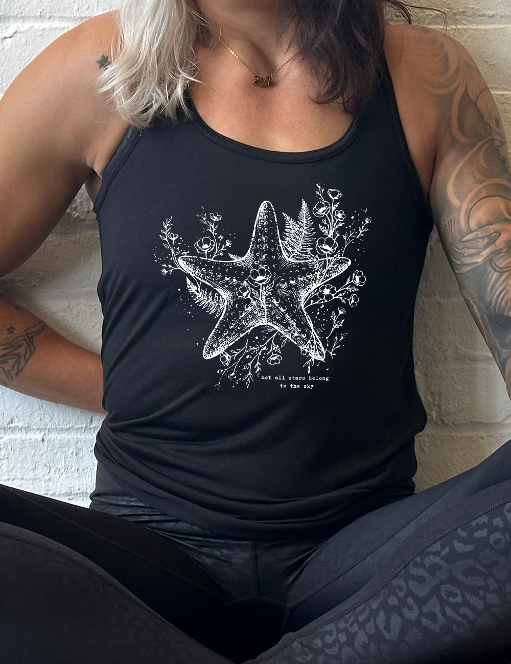 black racerback flowy tank with the graphic of a very detailed starfish among sea plants and flowers, with little bubbles, and the small words underneath it all that say "not all stars belong to the sky"