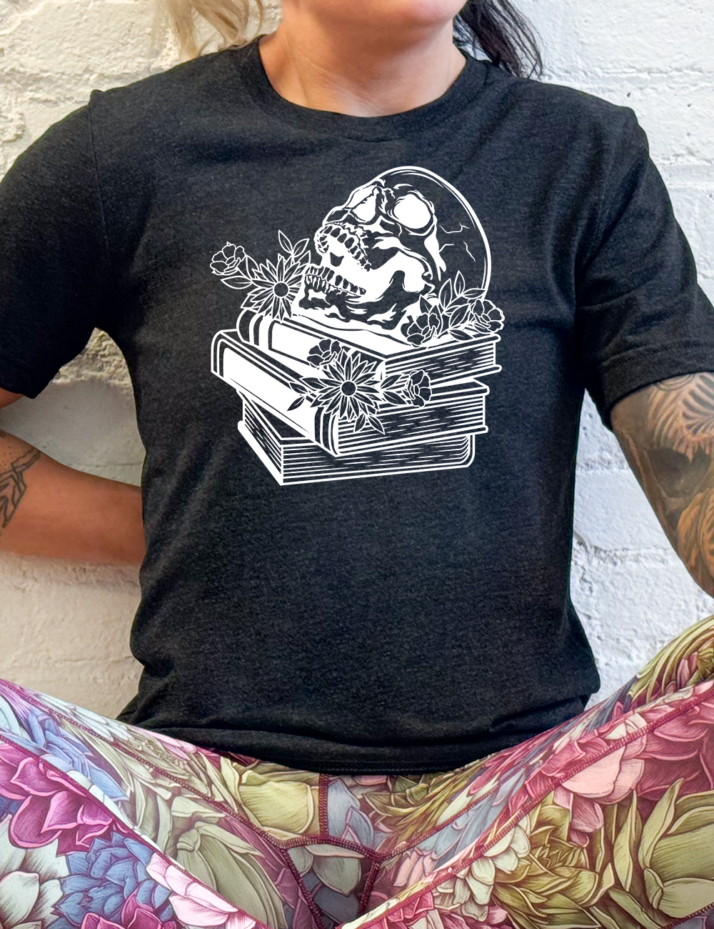 black unisex tee with the graphic of an open-mouthed skull atop a stack of books, with flowers