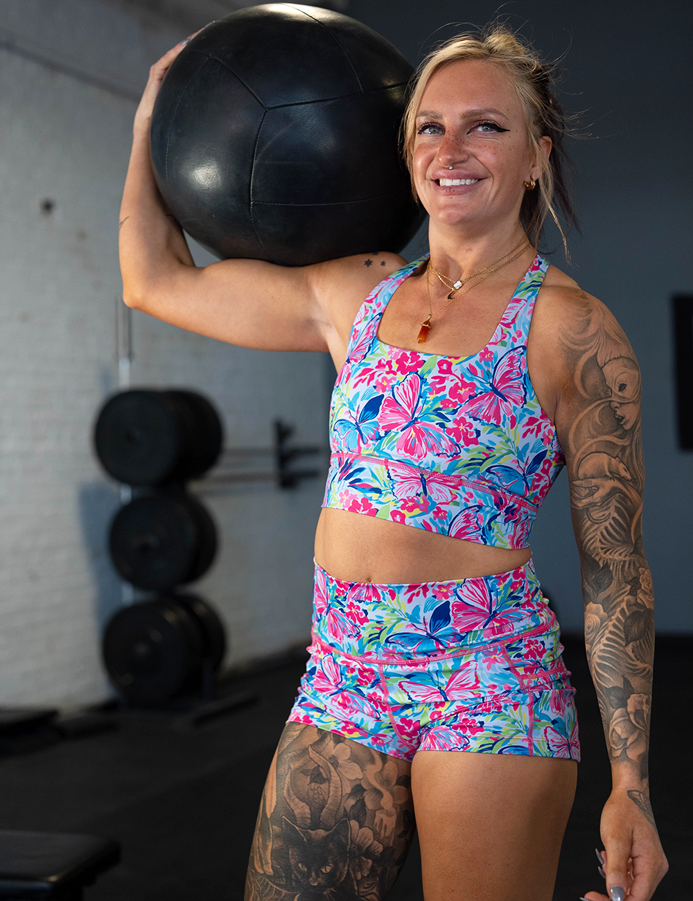 model wearing the pink and blue butterflies against a floral background shorts and bra in the gym lifting a medicine ball